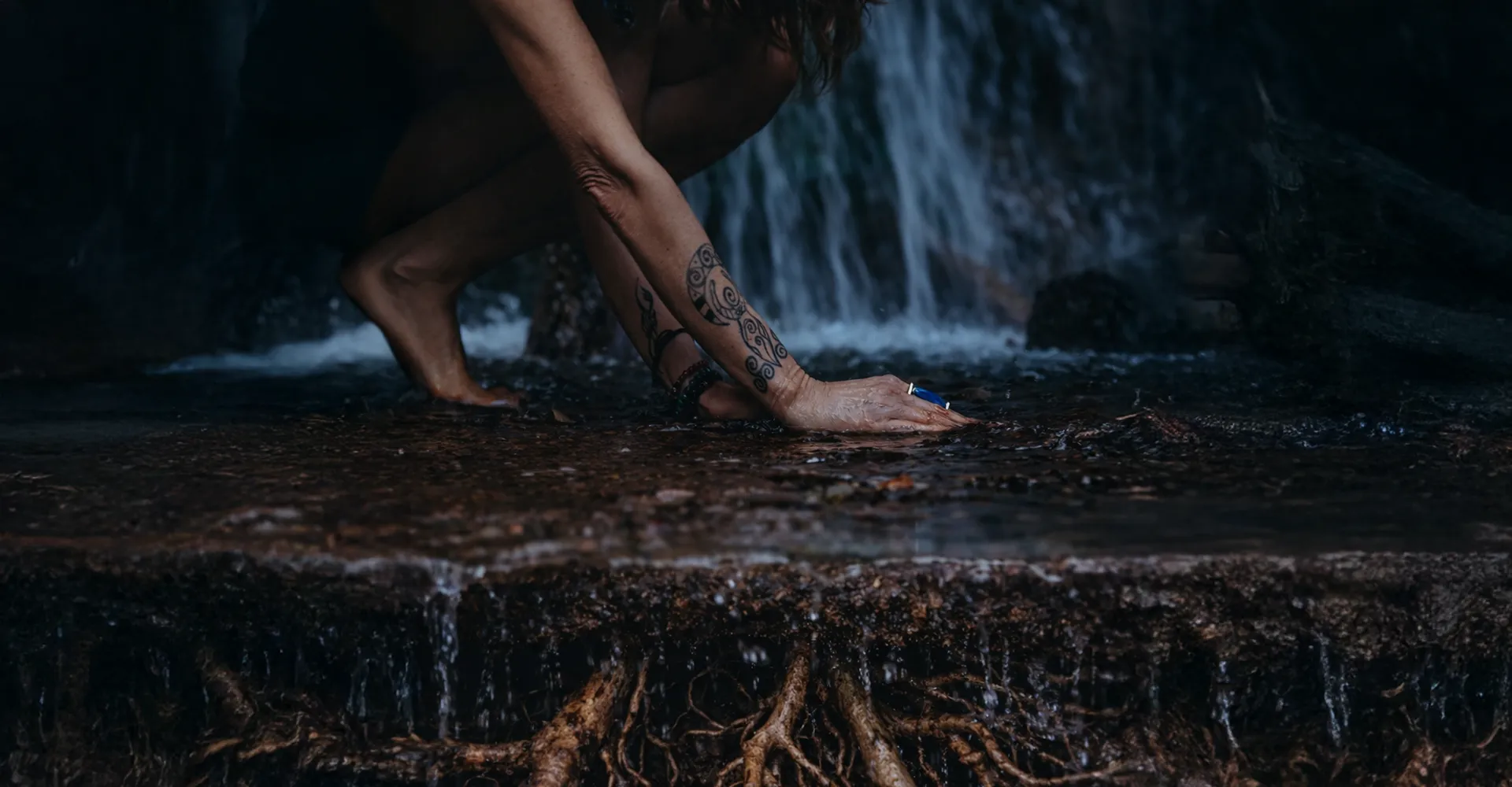 Una donna scalza, accovacciata su una roccia, immerge la mano nell'acqua di una piccola cascata